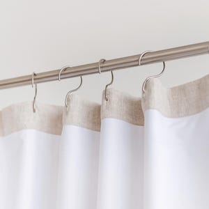 May include: White shower curtains with a neutral-colored border hang from a silver rod with curved hooks. The curtains are made of a smooth, opaque material, and the border has a textured appearance. The design is simple and modern, suitable for a bathroom.