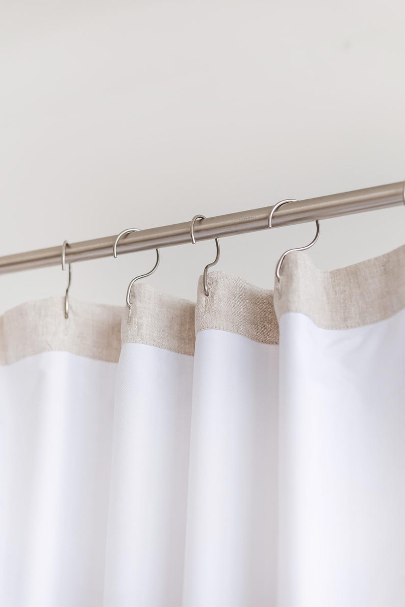 May include: White shower curtains with a neutral-colored border hang from a silver rod with curved hooks. The curtains are made of a smooth, opaque material, and the border has a textured appearance. The design is simple and modern, suitable for a bathroom.