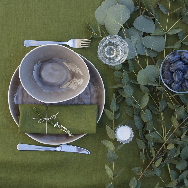 Pu&ograve; includere: Una mise en place con un piatto da portata in gres grigio, una ciotola in gres grigio pi&ugrave; piccola, un tovagliolo di lino, una forchetta d'argento, un coltello d'argento e una candela bianca. Il tavolo &egrave; coperto da una tovaglia di lino verde e circondato da rami di eucalipto.