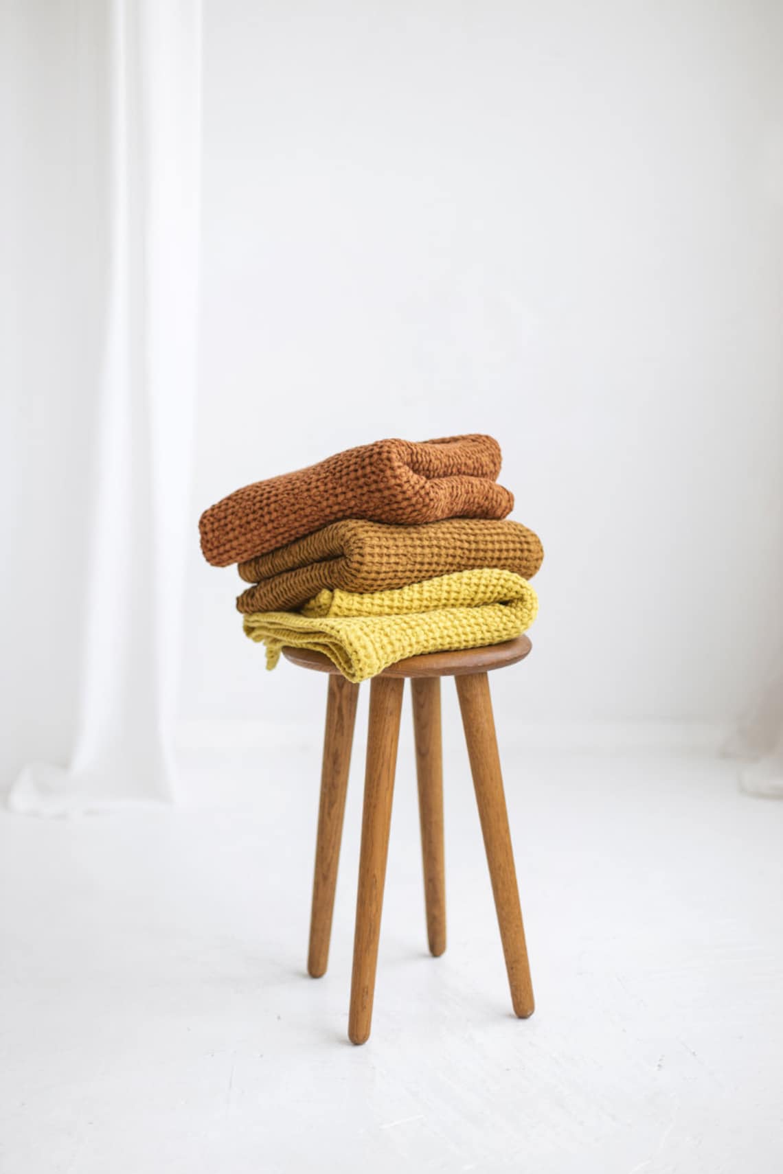 Three handmade linen waffle towels shown in different warm palette colors and displayed stacked on a wooden stool.