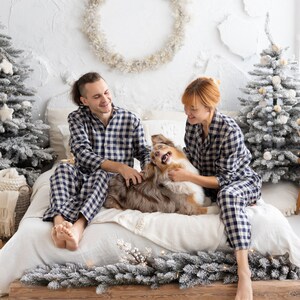 May include: A couple and a dog are on a bed, all wearing matching navy blue and white checkered pajamas. The dog is brown and white. The bed is decorated for the holidays with small Christmas trees and a garland.