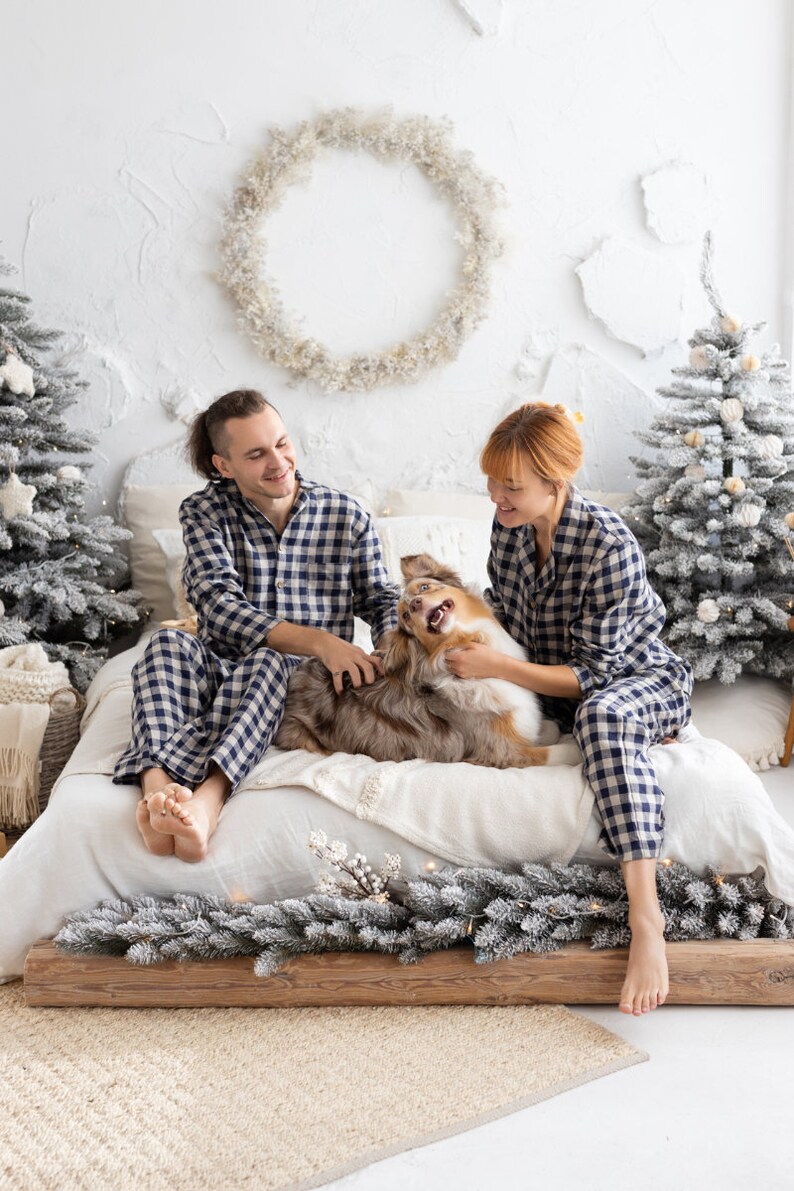 May include: A couple and a dog are on a bed, all wearing matching navy blue and white checkered pajamas. The dog is brown and white. The bed is decorated for the holidays with small Christmas trees and a garland.