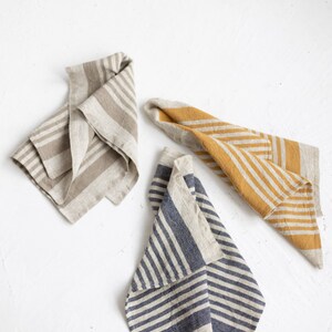 May include: Three linen kitchen towels folded in a triangular shape. The towels are striped with different colors: beige and brown, yellow and beige, and blue and beige.