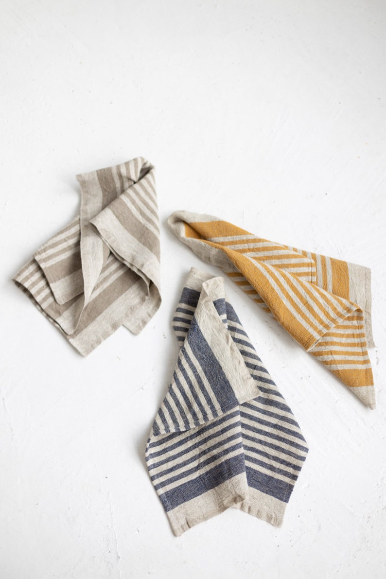 May include: Three linen kitchen towels folded in a triangular shape. The towels are striped with different colors: beige and brown, yellow and beige, and blue and beige.