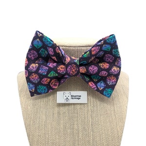 Puede incluir: Una pajarita negra con un patrón de dados coloridos. Los dados son en tonos de rosa, azul, verde y amarillo. La pajarita está en un maniquí marrón.