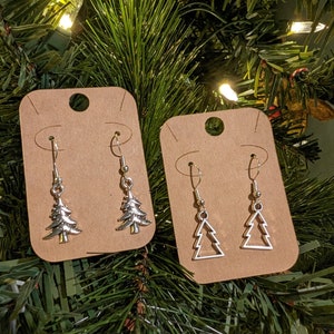 Può includere: Due paia di orecchini argentati a forma di albero di Natale su cartoncini marroni. Un paio presenta disegni di alberi pieni, l'altro contorni aperti. Gli orecchini sono appesi su rami verdi di albero di Natale.