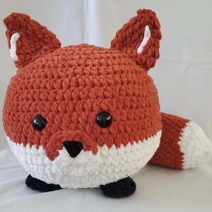 May include: A plush, round fox toy in shades of orange, white, and black. The fox has large, black eyes, a white muzzle, and a fluffy tail. The ears have white accents. The toy is made of a soft, textured material.