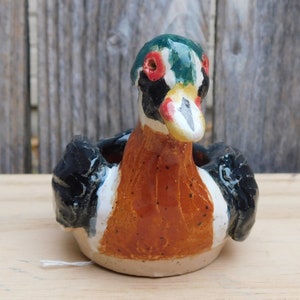 May include: A ceramic duck figurine with a colorful design. The duck has a red, white, and black head with a green top. The body is orange and white, with black wings. The figurine is sitting on a light-colored base.
