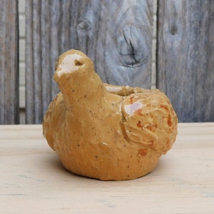 May include: A handcrafted, ceramic bird figurine in a warm, earthy tone. The bird has a rounded body, a small head, and a textured surface. It is a decorative item, likely for home decor.