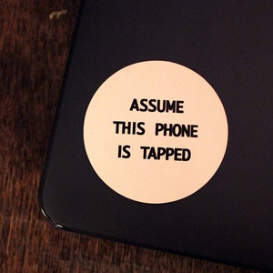 May include: A black laptop with a round, beige sticker that says "Assume this phone is tapped".