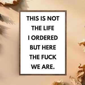 May include: A framed print with the text "THIS IS NOT THE LIFE I ORDERED BUT HERE THE FUCK WE ARE" in bold black letters on a white background. The frame is a dark brown colour.