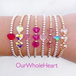 Könnte beinhalten: Ein Set aus sieben Armbändern mit goldenen und silbernen Perlen und Kristall-Herz-Anhängern in verschiedenen Farben, darunter Pink, Rot und Irisierend. Die Armbänder sind auf dem Handgelenk einer Person gestapelt. Der Text "OurWholeHeart" ist am unteren Rand des Bildes sichtbar.