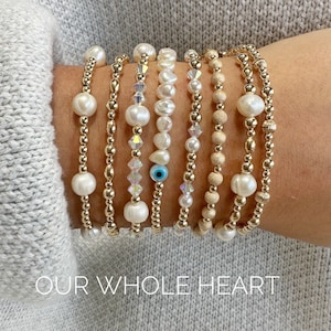 May include: A stack of gold bracelets with different beads, including pearls, crystals, and a blue evil eye charm. The bracelets are on a person's wrist and the text "OUR WHOLE HEART" is below the bracelets.
