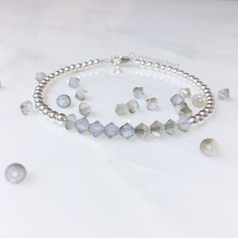 May include: A silver beaded bracelet with a row of clear iridescent glass beads. The bracelet is on a white surface with loose beads scattered around it. The text "OurWholeHeart" is visible in the image.