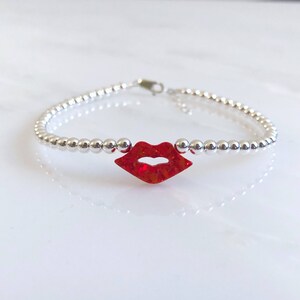 Puede incluir: Un brazalete de cuentas plateadas con un dije de labios rojos con brillo.