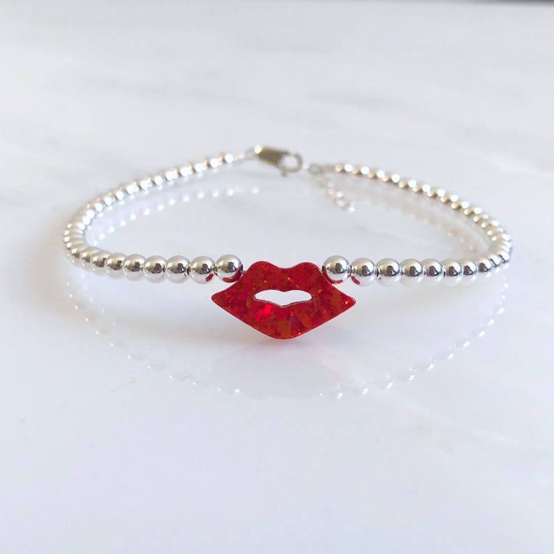 Puede incluir: Un brazalete de cuentas plateadas con un dije de labios rojos con brillo.