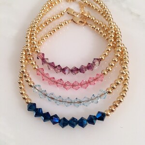 May include: Four gold beaded bracelets with crystal accents. The bracelets are stacked on top of each other, with the top bracelet featuring dark blue crystals, the next bracelet featuring light blue crystals, the next bracelet featuring pink crystals, and the bottom bracelet featuring purple crystals.