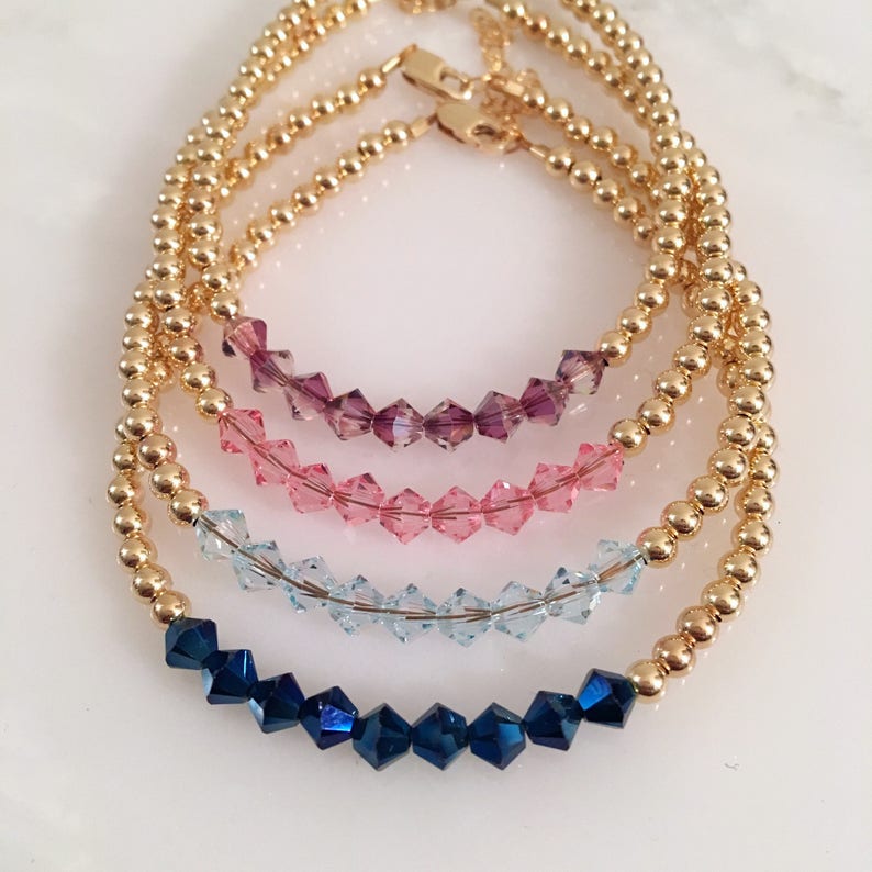 May include: Four gold beaded bracelets with crystal accents. The bracelets are stacked on top of each other, with the top bracelet featuring dark blue crystals, the next bracelet featuring light blue crystals, the next bracelet featuring pink crystals, and the bottom bracelet featuring purple crystals.