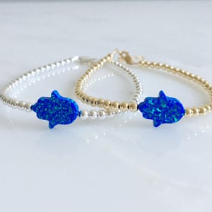 May include: Two bracelets with blue opal hamsa charms. One bracelet is silver and the other is gold.