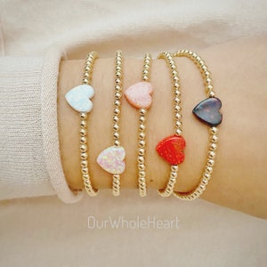 May include: Four gold beaded bracelets with heart-shaped charms in various colors, including white, pink, red, and black. The bracelets are stacked on a person's wrist. The text "OurWholeHeart" is visible below the bracelets.