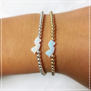 May include: Two beaded bracelets, one silver and one gold, each featuring a charm shaped like the state of New Jersey. The charms are white with an iridescent sheen. The bracelets are worn on a person's wrist.