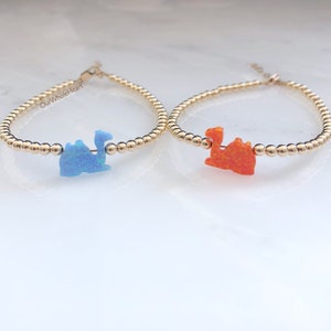 Peut inclure: Deux bracelets en perles dorées avec des breloques de chameau en opale. Un charme est bleu et l'autre est orange. Les bracelets sont sur une surface blanche. Le texte "OurWholeHeart" est visible sur le bracelet de gauche.