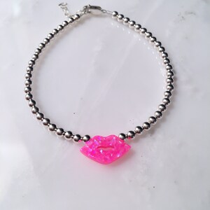 Puede incluir: Un brazalete de cuentas plateadas con un dije de labios de &oacute;palo rosa.