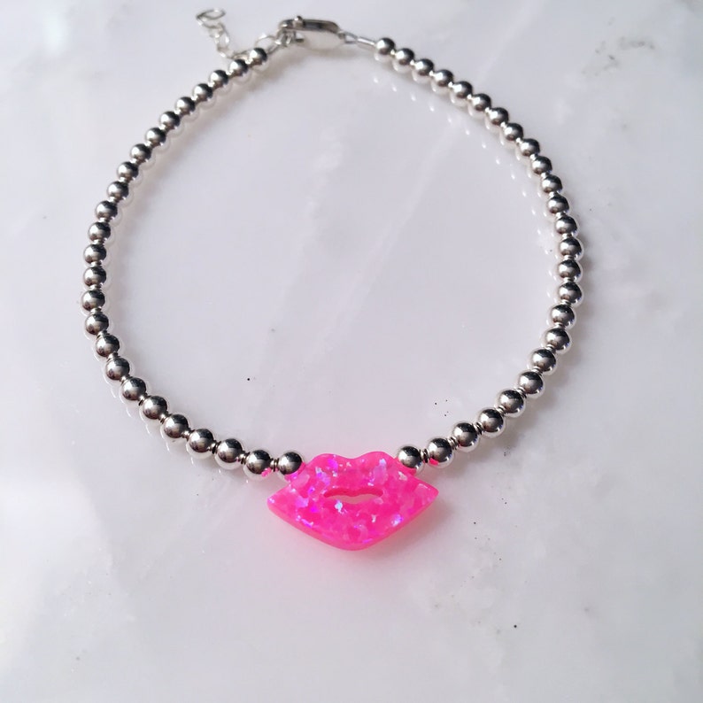 Puede incluir: Un brazalete de cuentas plateadas con un dije de labios de &oacute;palo rosa.