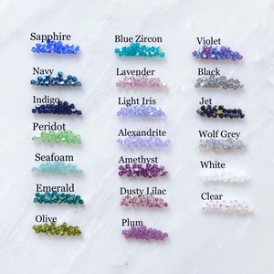 May include: A chart of 18 different colors of beads, each labeled with a color name. The colors include sapphire, blue zircon, violet, navy, lavender, black, indigo, light iris, jet, peridot, alexandrite, wolf grey, seafoam, amethyst, white, emerald, dusty lilac, clear, olive, and plum.