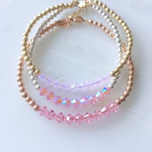 May include: Three delicate bracelets. The top bracelet is gold, the middle is silver, and the bottom is rose gold. Each bracelet features small, round beads. The middle and bottom bracelets have pink crystal accents.