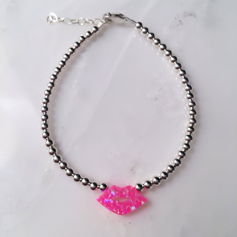 Puede incluir: Un brazalete de cuentas plateadas con un dije de labios de &oacute;palo rosa.