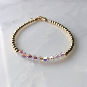 May include: A gold beaded bracelet with iridescent crystal beads. The bracelet is made of small gold beads and features a row of clear crystal beads with an iridescent finish.