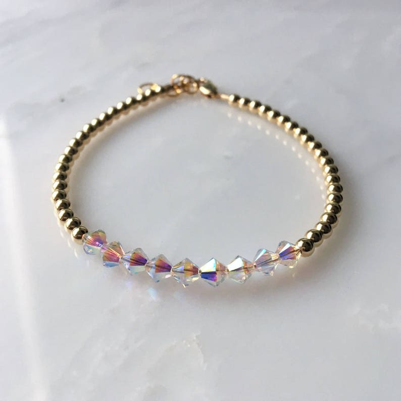 May include: A gold beaded bracelet with iridescent crystal beads. The bracelet is made of small gold beads and features a row of clear crystal beads with an iridescent finish.