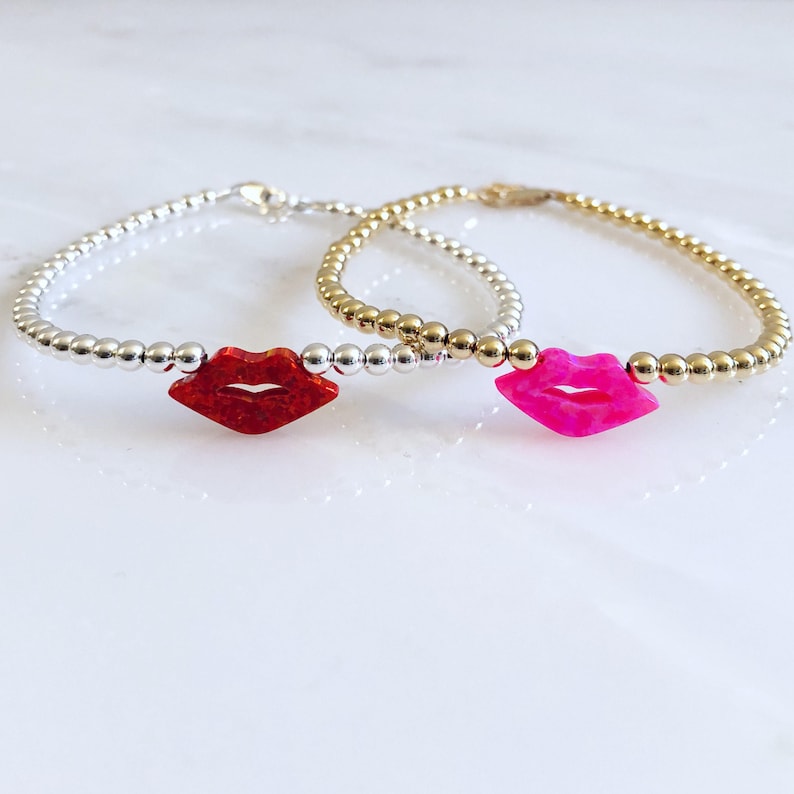 Puede incluir: Dos pulseras delicadas con cuentas plateadas y doradas, cada una con un dije en forma de labio rojo y rosa. Los dijes son de &oacute;palo.