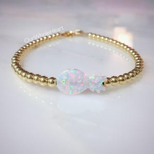 May include: A gold beaded bracelet with a white opal pineapple charm. The bracelet is delicate and feminine, perfect for adding a touch of whimsy to any outfit.