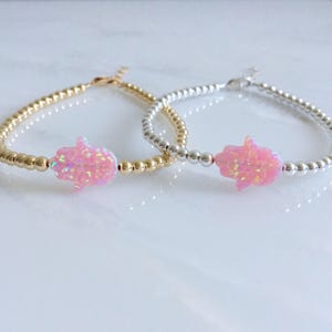 May include: Two delicate bracelets with a pink opal hamsa charm. One bracelet is gold and the other is silver.