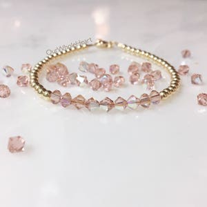 May include: A delicate gold bracelet with a row of pink crystal beads. The bracelet is on a white surface with loose pink crystal beads scattered around it.