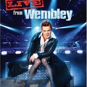 Peut inclure: Un comédien portant une veste en cuir noire et des bas résille noirs se produit en direct sur scène. La scène est éclairée par des projecteurs et l'arrière-plan est une grande foule de personnes. Le texte "LIVE from Wembley" est affiché au-dessus du comédien.