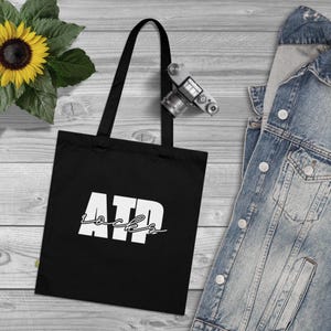 May include: Black tote bag with white text that reads "ATP Rocks". The bag is on a wooden surface with a denim jacket and a camera.