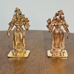 May include: Two gold-colored statues of a deity, each standing on a square base. The statues have intricate details, including ornate headdresses and multiple arms. They are positioned on a wooden surface.