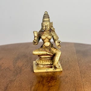 May include: A small, golden-colored statue of a seated figure with ornate headdress and jewelry. The figure holds an object in one hand and is seated on a detailed pedestal. The statue has a weathered appearance, suggesting age or a vintage aesthetic.