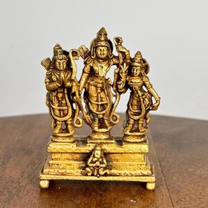 Puede incluir: Una estatua dorada que representa tres figuras sobre una base rectangular. Las figuras tienen ropa y tocados detallados. La estatua es probablemente un objeto religioso o decorativo.