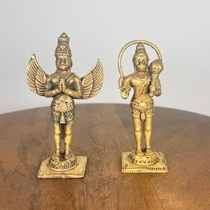 Peut inclure: Deux statues dorées de divinités. L'une des figures a des ailes déployées et les mains jointes. L'autre figure tient un bâton et a un halo. Les deux statues sont sur des bases carrées et sont posées sur une surface en bois.