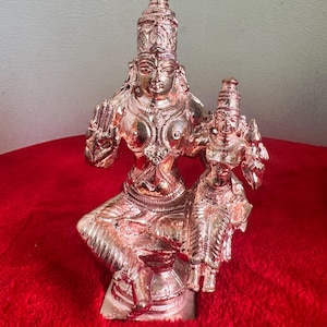 Può includere: Una statua dettagliata color bronzo di una divinità seduta che tiene una figura più piccola. La figura più grande è adornata con gioielli intricati e un'alta acconciatura. La statua è posta su uno sfondo rosso, mostrando l'artigianato dettagliato della scultura religiosa.