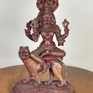 Op de afbeelding: Een bronzen beeld van een godheid die op een leeuw rijdt. De figuur heeft meerdere armen en draagt een kroon. Het beeld heeft rode accenten en staat op een decoratieve basis. De algehele stijl doet denken aan oosterse religieuze kunst.