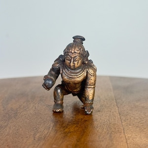 Può includere: Una piccola statuetta color bronzo raffigurante una figura inginocchiata con lineamenti dettagliati e copricapo ornato. La statua è posizionata su una superficie di legno, mettendo in risalto il suo design e la sua maestria. La figura sembra essere un manufatto religioso o culturale.