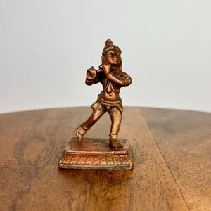 Könnte beinhalten: Eine bronzefarbene Figur einer Person, die Flöte spielt. Die Figur steht auf einem rechteckigen Sockel mit aufwendigen Details. Die Figur ist in einer dynamischen Haltung dargestellt, mit einem ausgestreckten Bein. Die Gesamtästhetik ist detailliert und künstlerisch.