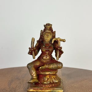 Peut inclure: Petite statue en bronze détaillée d'une divinité assise à plusieurs bras, ornée de sculptures complexes. La statue présente une finition dorée avec des accents rouges et bruns, et est montée sur une base carrée. La divinité est représentée avec une couronne et tient des objets dans ses mains.