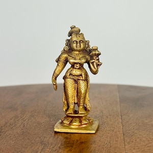May include: A small, gold-colored statue of a female figure, likely a deity, standing on a square base. The statue has intricate details, including ornate clothing and a headdress. The figure is holding an object in one hand.