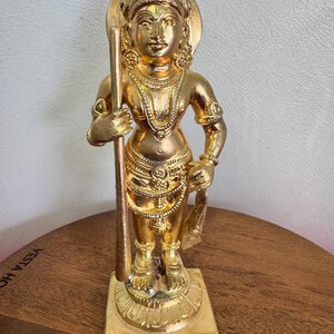 May include: A gold-coloured statue of a deity, standing upright and holding a staff. The statue is adorned with intricate jewellery and a halo. The base is square and the statue is on a wooden surface.